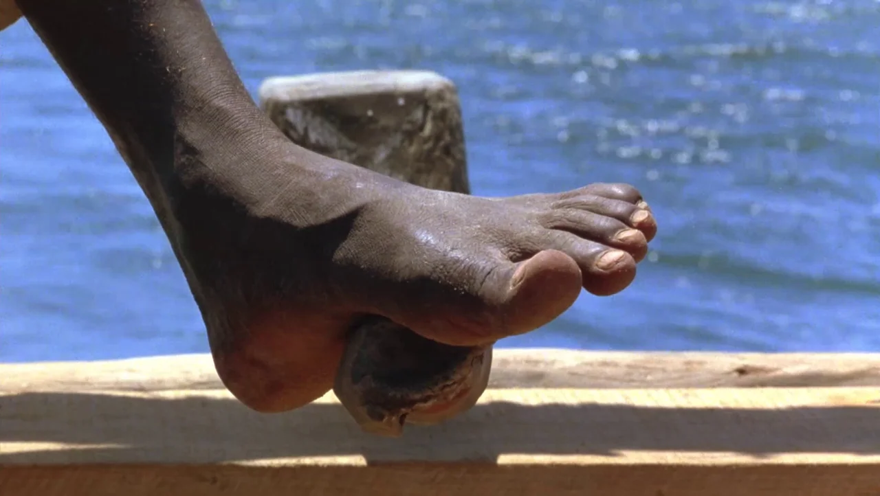 Mountains of the Moon (1990) movie still: closeup — A wooden pier structure sits above rippling blue water under a bright sun. A bare foot rests on…