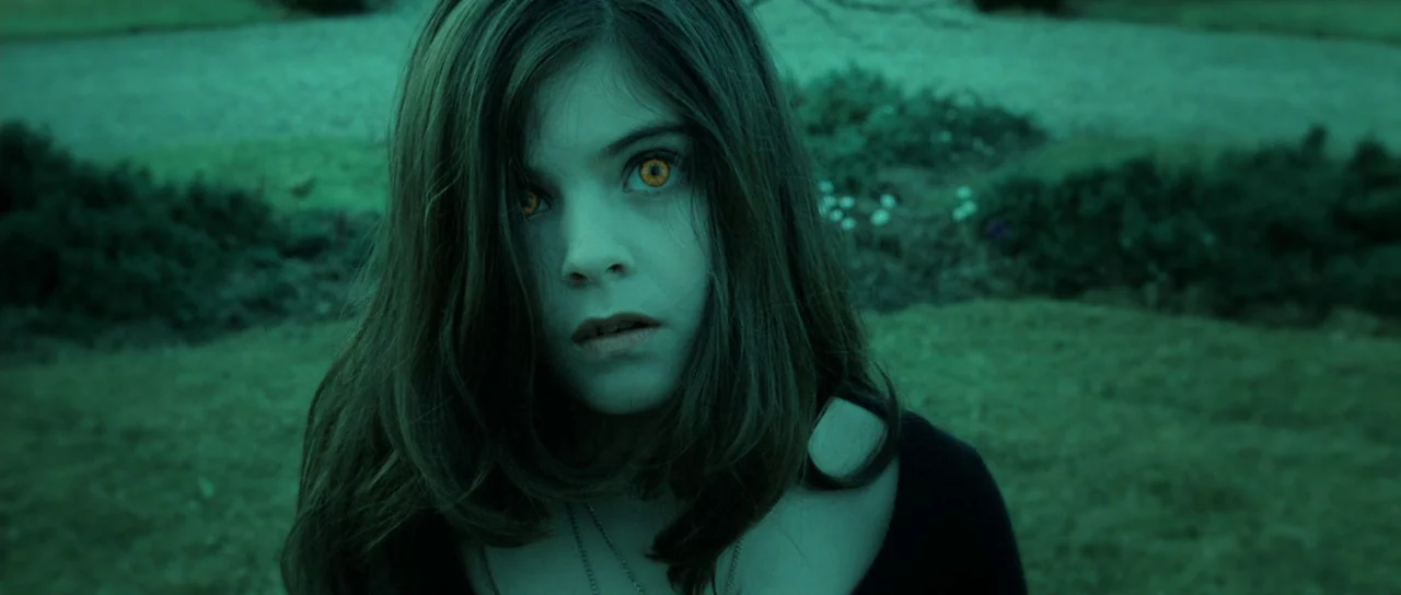 Daybreakers (2009) movie still: closeup — A dark, grassy expanse is visible in the soft green glow of night. A young girl with bright amber eyes…