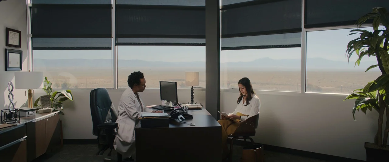 Intrusion (2021) movie still: wide — A modern medical office overlooks a wide, dry desert landscape through expansive windows. A doctor sits at a…