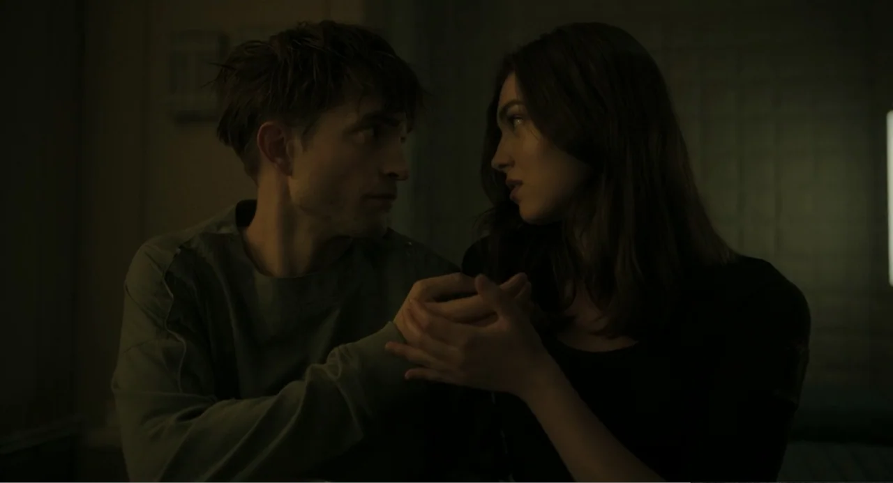 Mickey 17 (2025) movie still: medium — A dimly lit room with tiled walls and muted green tones. A man in a hospital gown holds hands with a woman.