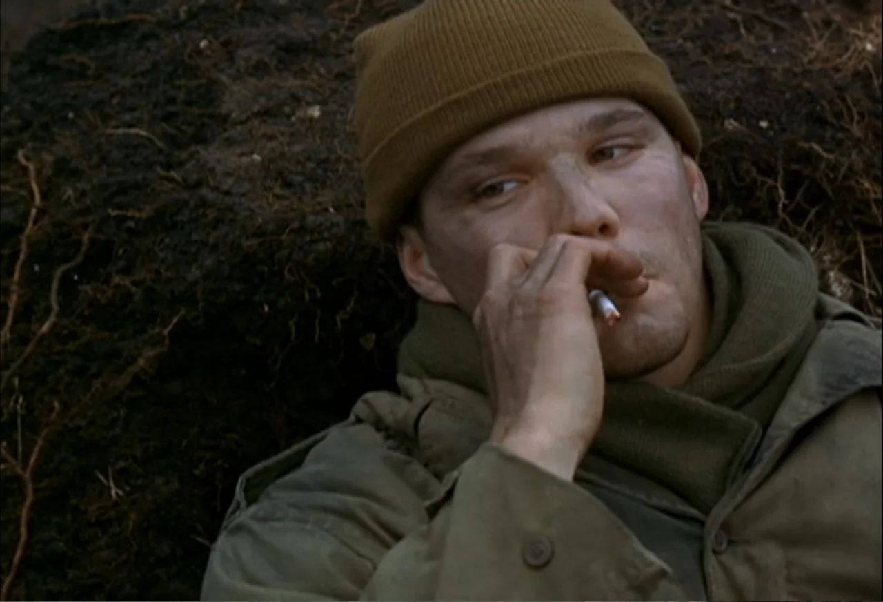 When Trumpets Fade (1998) movie still: closeup — A soldier lies in a mud-caked dirt trench outdoors. The man in military gear smokes a cigarette…
