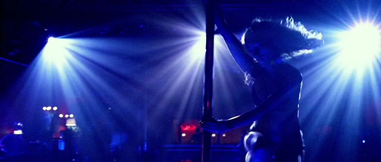 Too Late (2015) movie still: medium — A dark venue with intense blue backlighting focused on a dancer. The dancer holds onto a central metal pole in…
