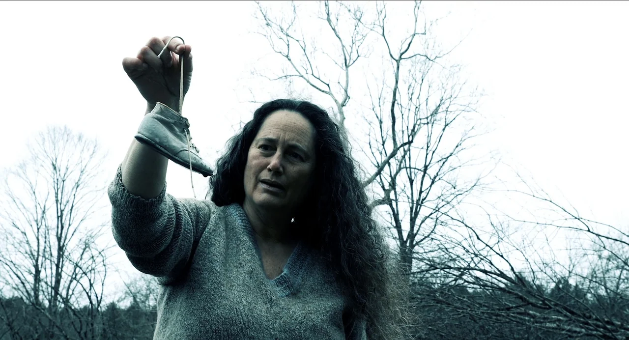 Where The Devil Roams (2023) movie still: medium — An outdoor area with leafless, barren trees against a bright, overcast white sky. A woman in a…