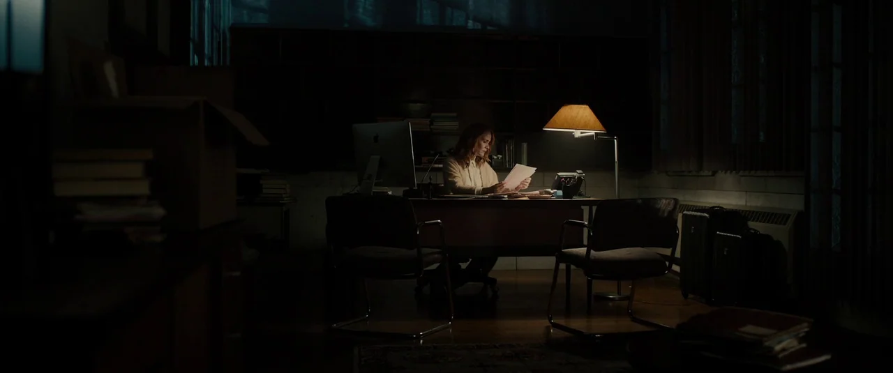 Glass (2019) movie still: wide — A dimly lit room with dark walls features a lone person working at a desk. A single table lamp illuminates the desk…