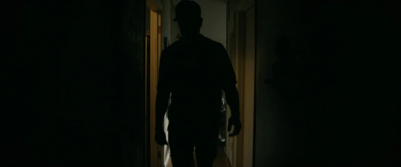 One Fast Move (2024) movie still: medium — A dark hallway connects two rooms in a residential space. A person walks forward in silhouette against…