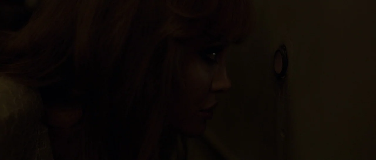 By The Sea (2015) movie still: closeup — A dimly lit indoor space with a plain, dark-colored wall surface. A woman leans forward, positioning her…