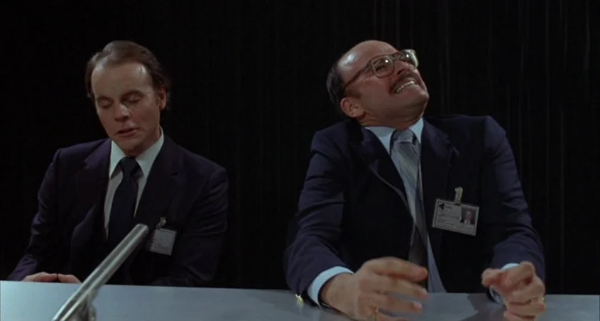 Scanners (1981) movie still: medium — Two men in business suits sit behind a desk against a plain black curtain. The man on the right looks toward…