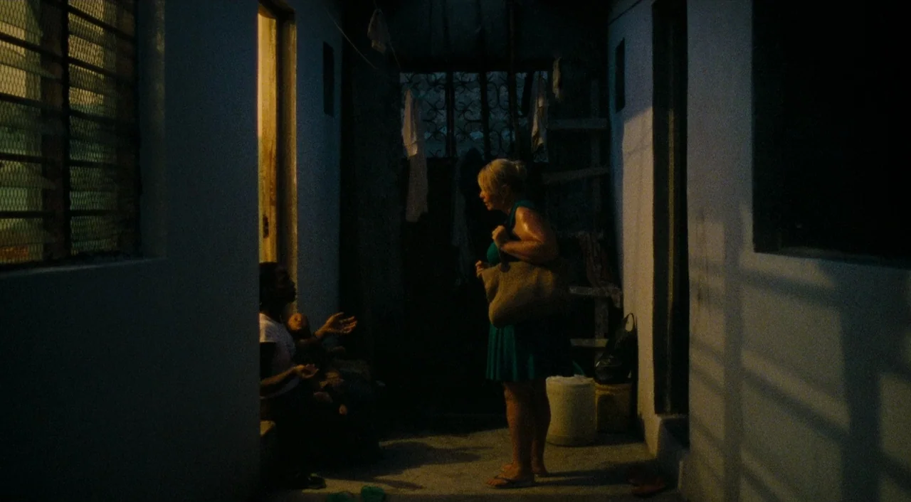 Paradise: Love (2012) movie still: fullbody — A narrow residential corridor at night with white walls and hanging laundry items visible. A woman in…