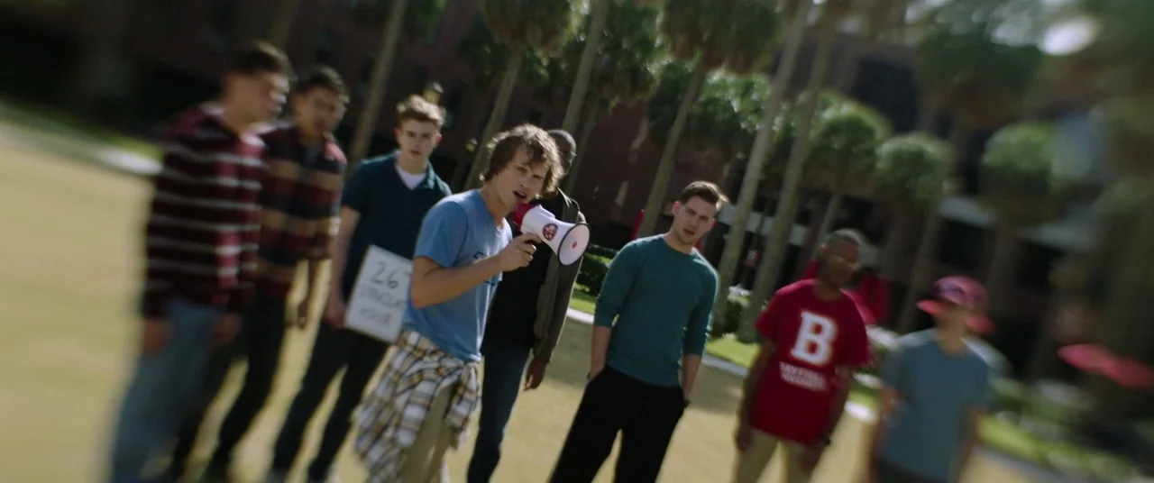 Happy Death Day (2017) movie still: wide — A group of young men stands on a grassy area in front of palm trees. One man holds a megaphone while…