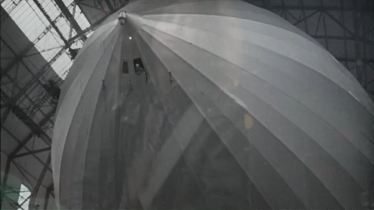 Becoming Led Zeppelin (2025) movie still: establishing — A large airship interior structure is housed inside a metallic industrial hangar building.…