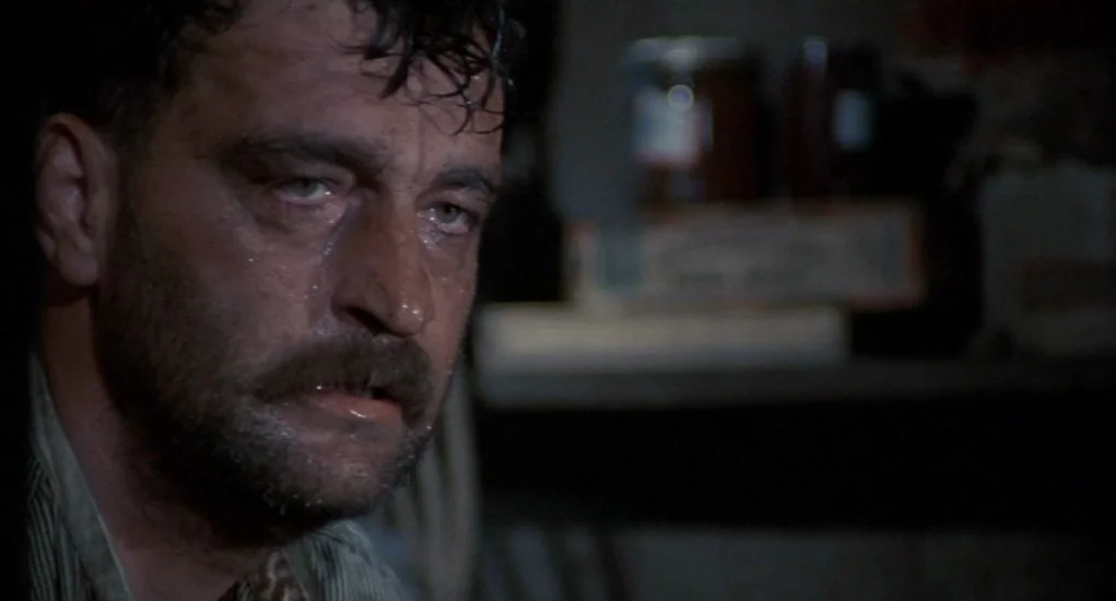 The Other (1972) movie still: closeup — A dark, cramped space contains shelves filled with jars in the soft background blur. A sweating man with a…