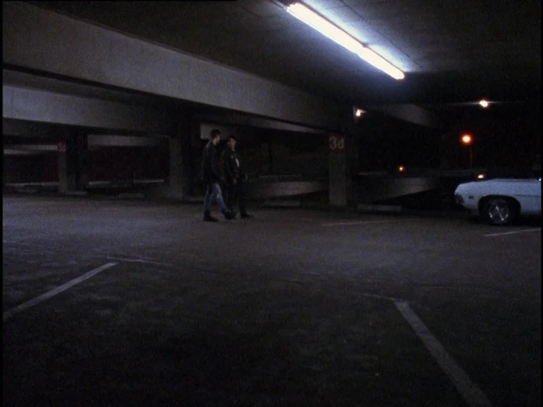 Totally F***ed Up (1993) movie still: wide — A dark, low-ceilinged parking structure made of concrete with visible structural columns. Two men walk…