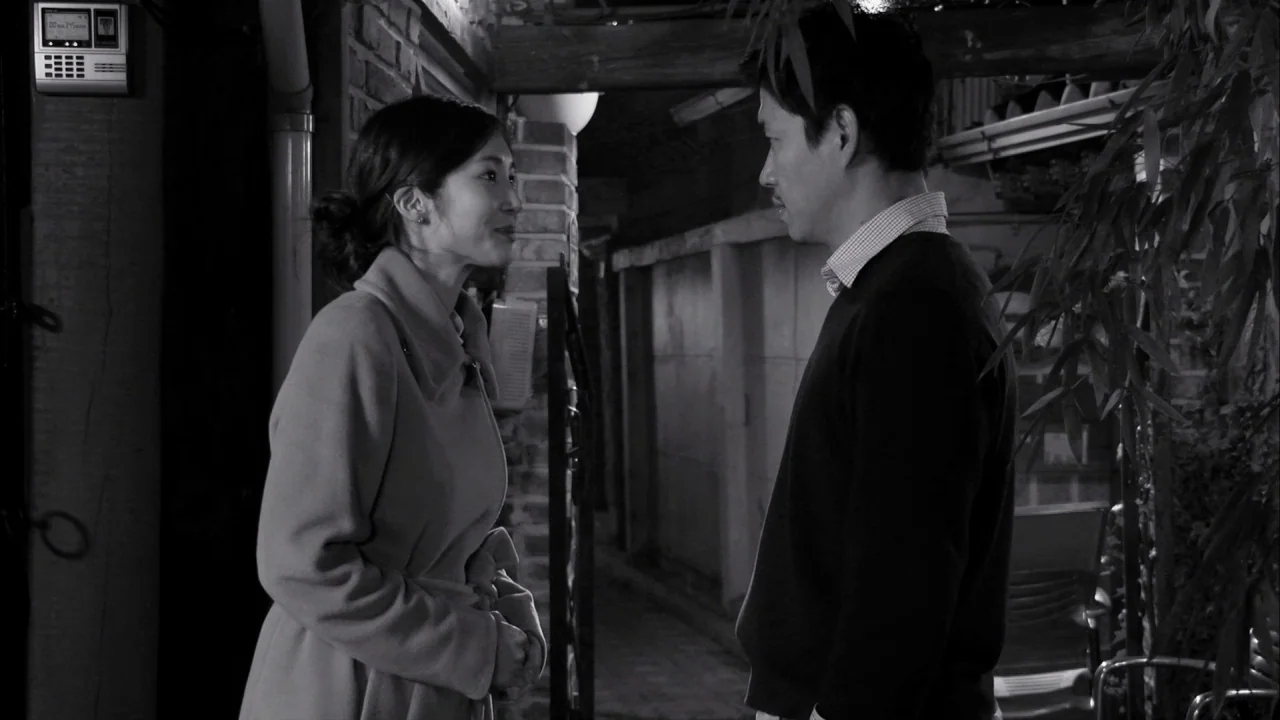 The Day He Arrives (2018) movie still: medium — An alleyway with brick walls at night provides a backdrop for two people. A man and woman stand…
