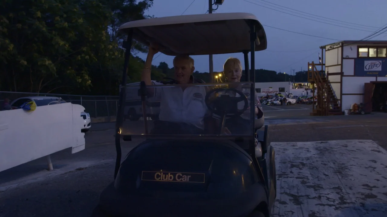 The Last Race (2022) movie still: medium — An outdoor paved area at a racing track features a dark golf cart. An elderly couple sits inside the golf…