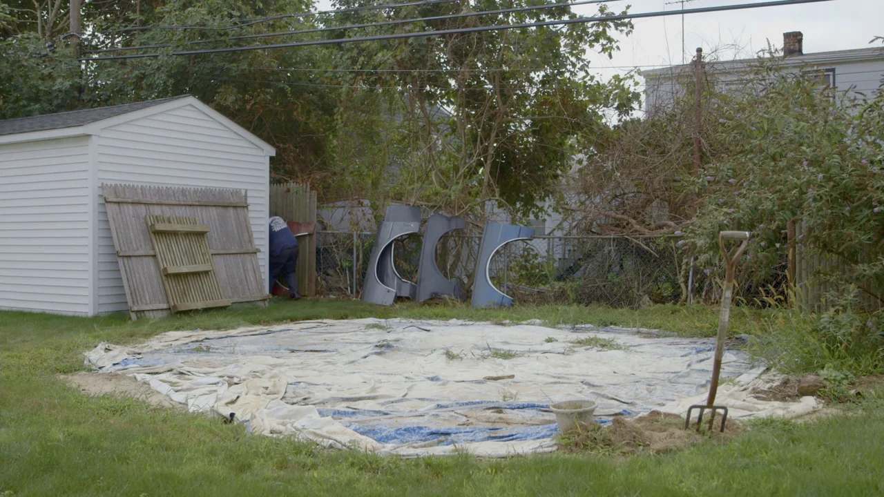 The Last Race (2022) movie still: wide — A suburban backyard features a white shed, a chain-link fence, and a tarp. A man stands behind the fence…