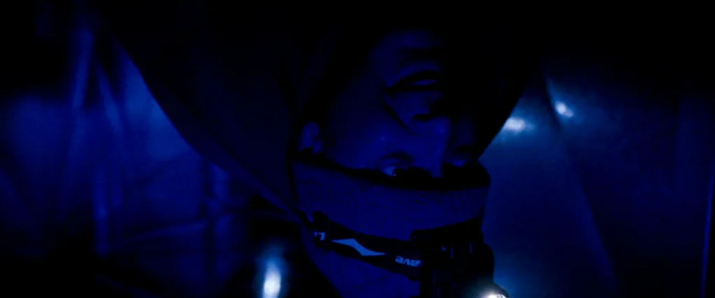 Ocean’s Thirteen (2007) movie still: closeup — A man looks down from an inverted perspective inside a dark confined space. He wears a headlamp that…