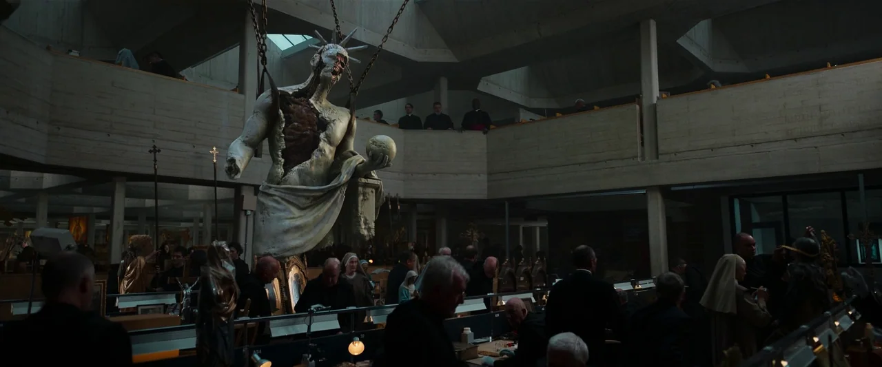 The Pope’s Exorcist (2023) movie still: wide — A large, modern, brutalist-style hall contains numerous individual work stations arranged in rows. A…