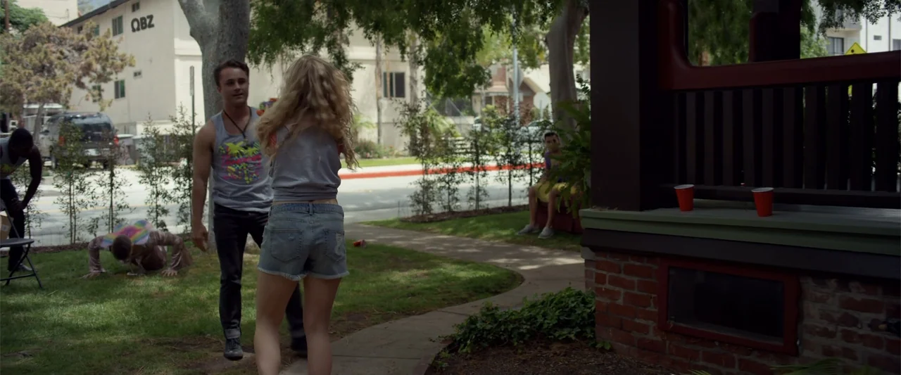 Init!ation (2021) movie still: medium — A residential front yard features green grass, a paved path, and mature trees. A man and a woman stand on…