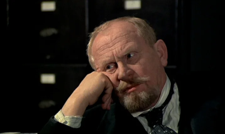 The Serpent’s Egg (1977) movie still: closeup — A dark, dimly lit archive room with black file drawers in the blurred background. An elderly man…