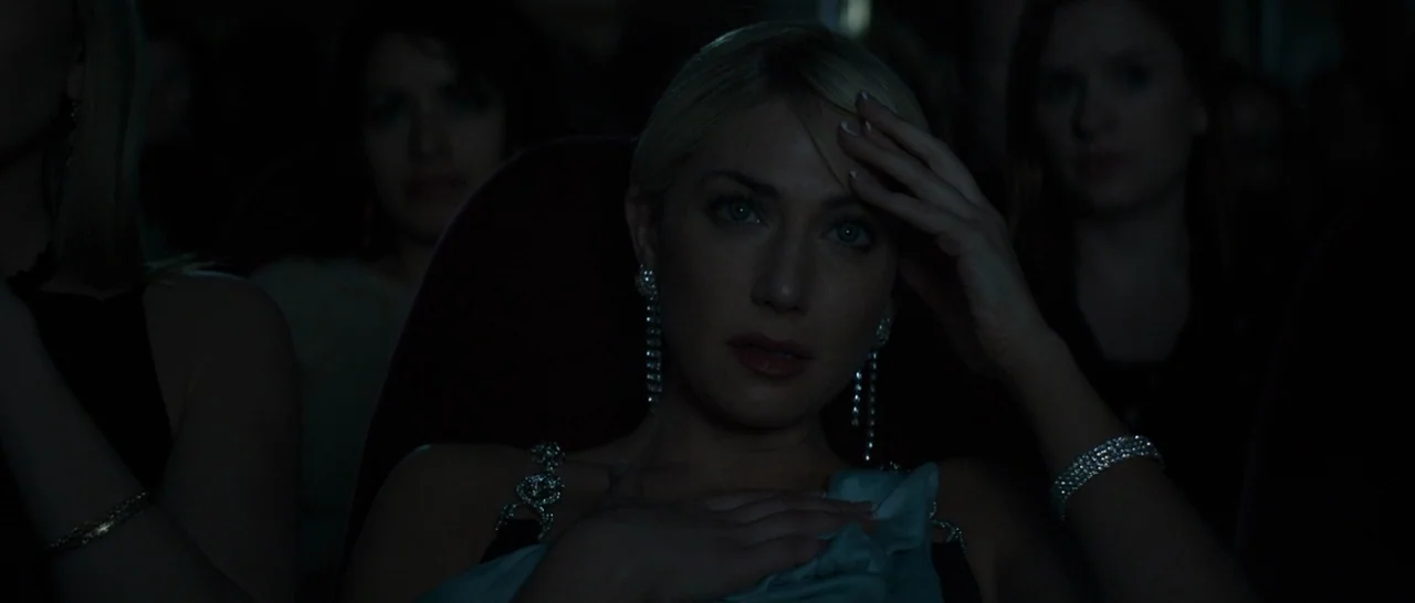 The Disaster Artist (2017) movie still: closeup — A dimly lit interior space with several people seated in plush dark chairs. A blonde woman with…