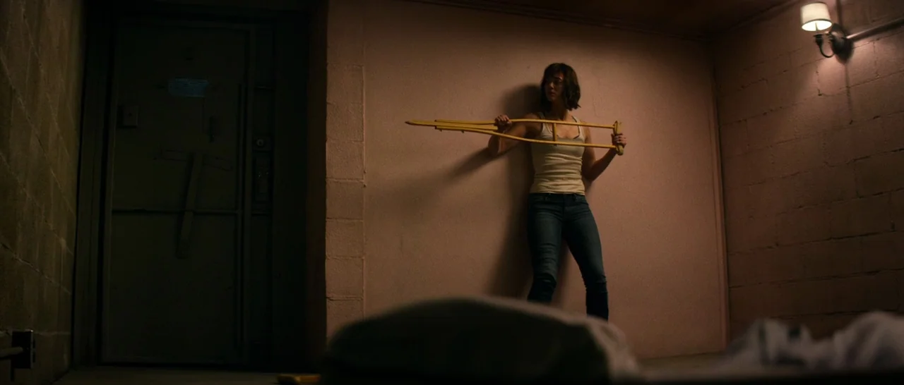 10 Cloverfield Lane (2016) movie still: fullbody — A concrete bunker with smooth beige walls and a heavy metal door. A young woman stands against…
