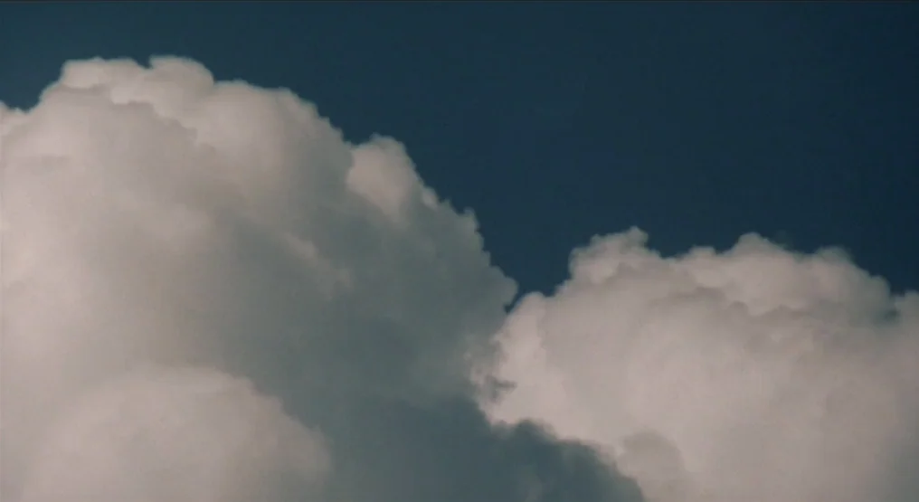 Rhapsody in August (1991) movie still: establishing — Dense, rounded cumulus clouds fill most of the frame against a clear sky. The sun shines…