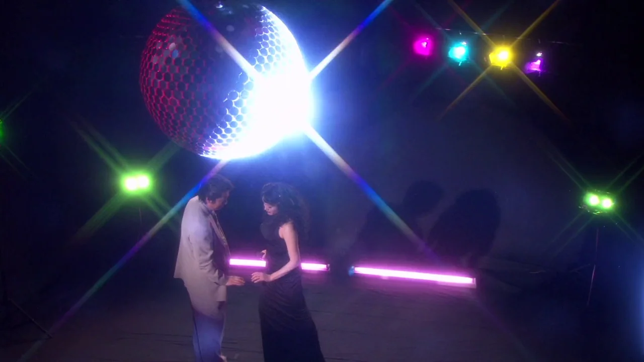 The Happiness of the Katakuris (2001) movie still: fullbody — A dark, expansive room is illuminated by colorful neon tubes and a large disco ball. A…