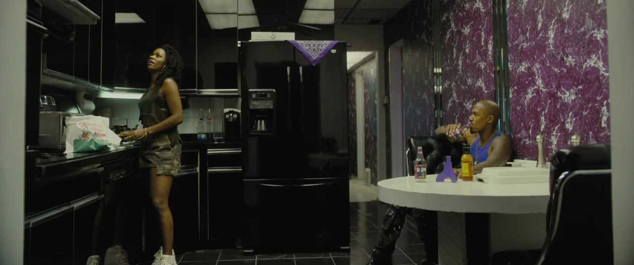 Chi-raq (2015) movie still: wide — A modern kitchen features black lacquer cabinets and high-contrast purple faux-marble patterned walls. A woman…