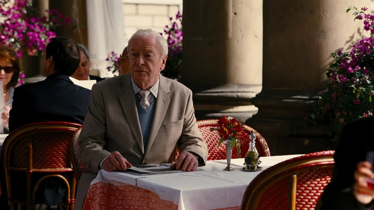 The Dark Knight Rises (2012) movie still: medium — An outdoor cafe terrace features stone pillars and bright pink bougainvillea in the background.…
