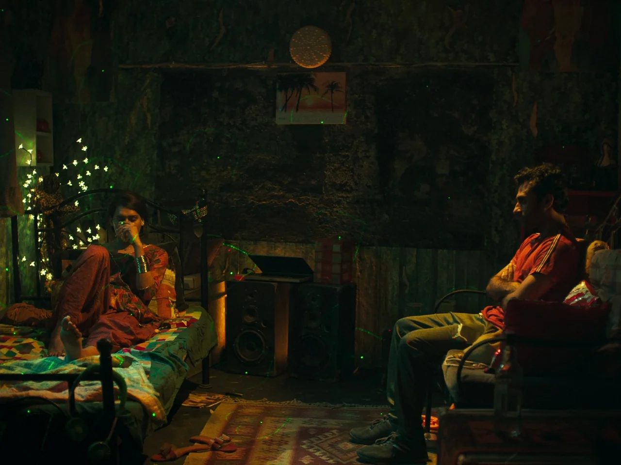 Joyland (2022) movie still: medium — A dimly lit room is illuminated by scattered green laser light patterns. A man sits facing a woman who is…