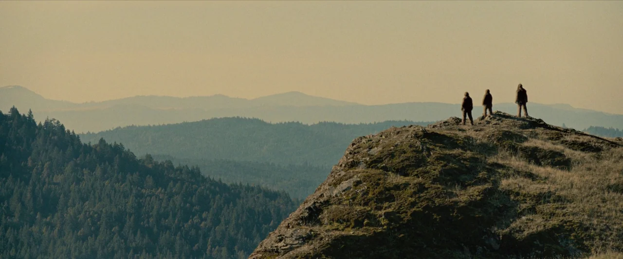 Sasquatch Sunset (2024) movie still: wide — A rugged mountain ridgeline overlooks a vast, hazy forest under a pale sky. Three people stand at the…