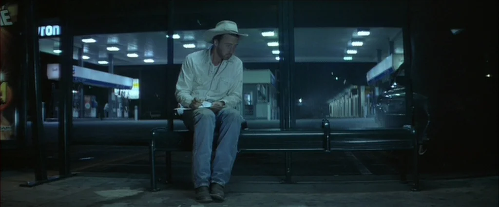 Down In The Valley (2005) movie still: fullbody — A man sits alone on a dark bench at a bus stop at night. Behind him are the bright, glowing lights…