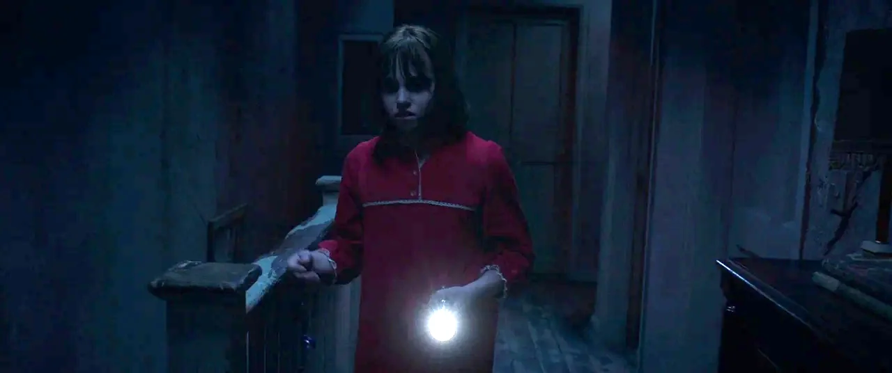The Conjuring 2 (2016) movie still: medium — A dark house interior with a wooden banister at night. A young girl in a red nightgown moves through…