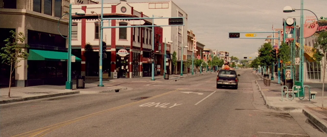 Carriers (2009) movie still: wide — An empty city street is lined with multi-story brick commercial buildings and green awnings. A single dark SUV…