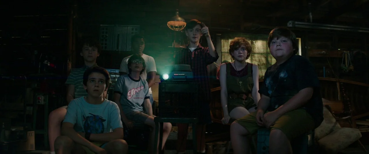 It (2017) movie still: wide — A dimly lit garage filled with clutter and wooden structures during the nighttime. Six adolescents are gathered around…