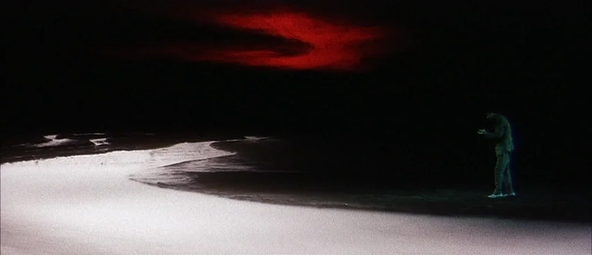 Jigoku (1960) movie still: wide — A dark, empty beach at night is illuminated by a narrow strip of white light. A solitary man stands on the sand…