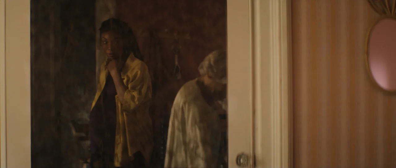 The Front Room (2024) movie still: medium — A dimly lit indoor corridor with striped wallpaper features a woman standing by a doorway. A second…