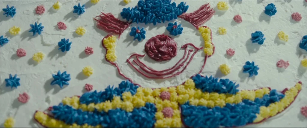 Clown (2014) movie still: closeup — A rectangular cake is covered in smooth white frosting with a clown drawing. The clown face is made of piped…