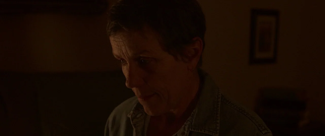 Nomadland (2020) movie still: closeup — A dimly lit interior room at night with dark brown walls. An older woman with short hair looks down while…