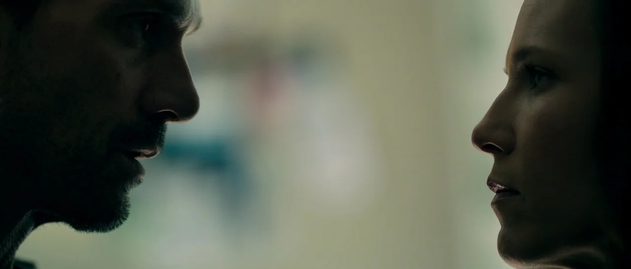 The Purge: Anarchy (2014) movie still: closeup — A close up shows two individuals facing each other from profile in a dim room. The light hits the…