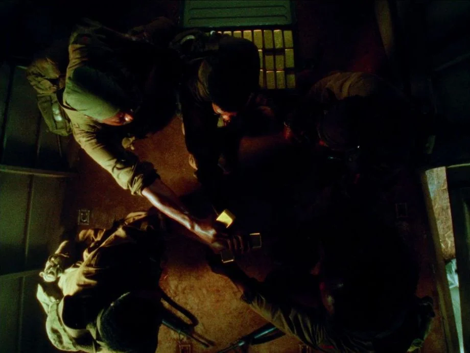 Da 5 Bloods (2020) movie still: wide — The interior of a darkened cargo hold displays heavy metal flooring and stacked crates. Four soldiers in…