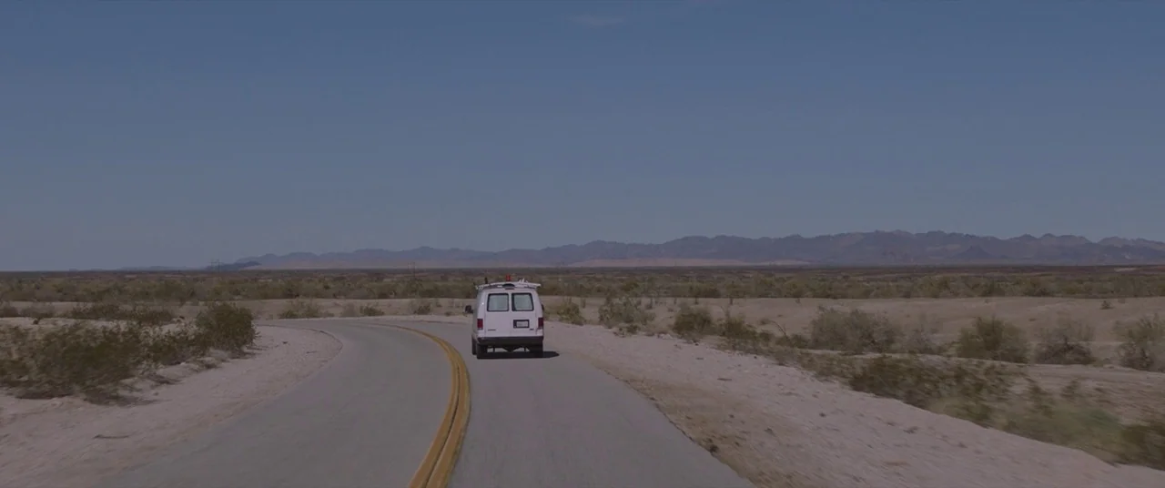 Nomadland (2020) movie still: wide — A white cargo van travels down a desolate paved road through a barren desert. Distant rolling mountains sit…