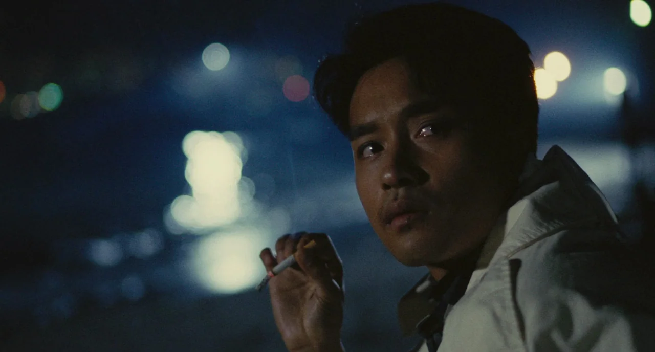 A Better Tomorrow (1986) movie still: closeup — A dark night at the beach features blurred city lights reflecting on the water. A man in a light…