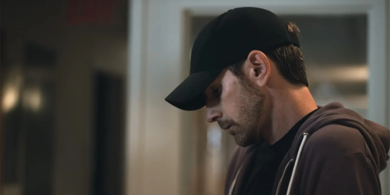 Equal Standard (2021) movie still: closeup — An interior room with a blurred white doorway visible in the background. A man in a baseball cap and…