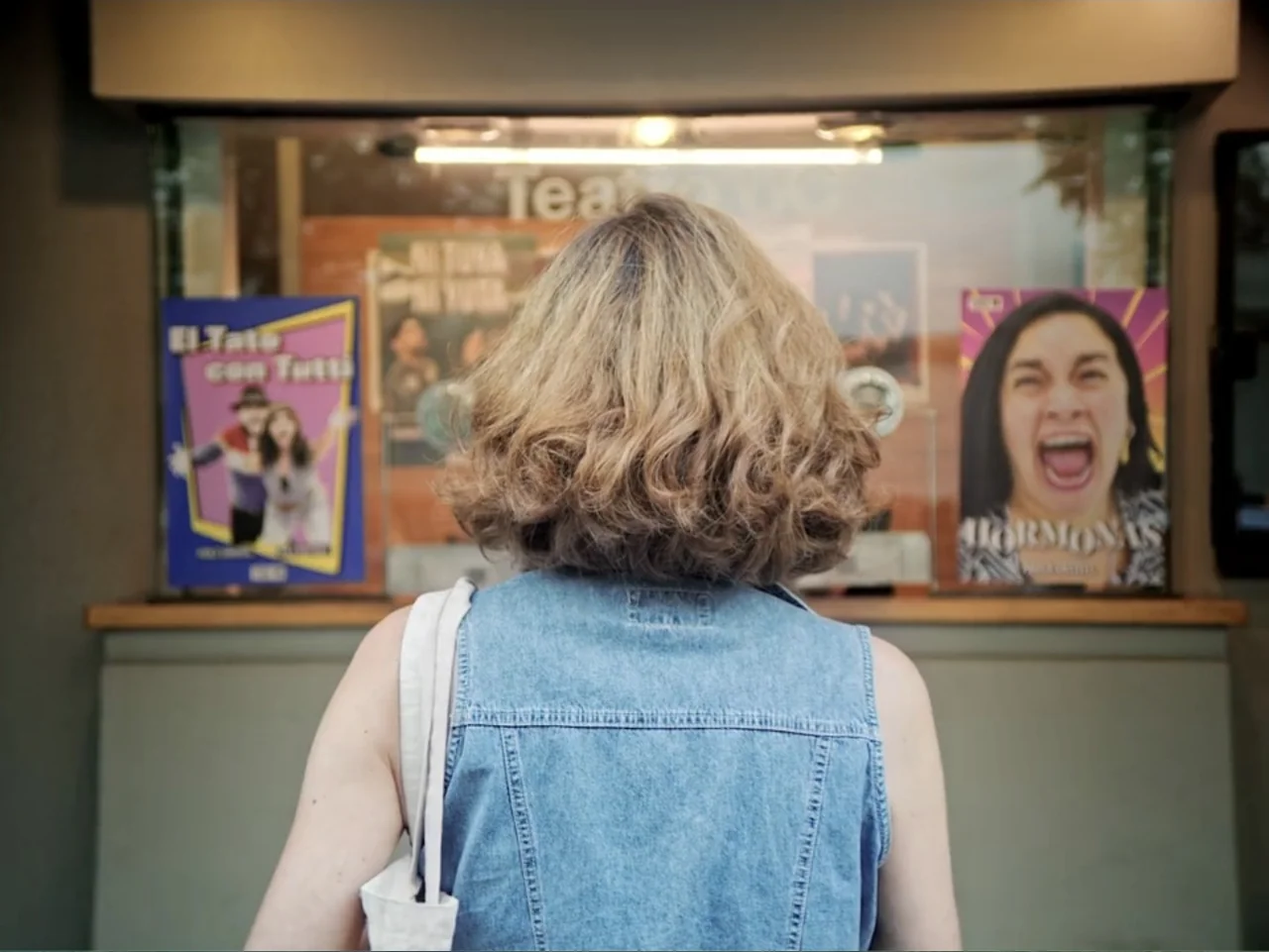 History and Geography (2024) movie still: medium — A woman stands before a glass ticket booth display featuring various theater event posters. She…
