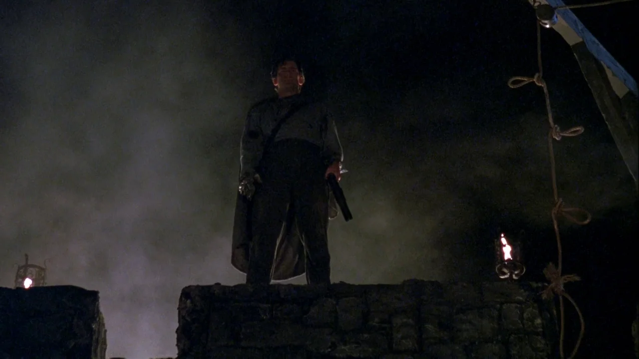 Army of Darkness (1992) movie still: fullbody — A man stands atop stone battlements at night silhouetted against a dark, foggy sky. He holds a…