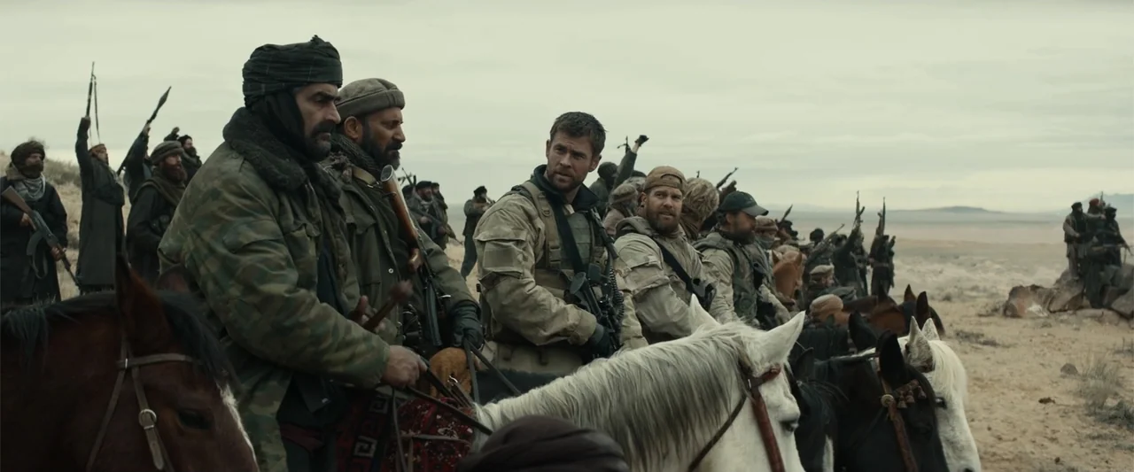 12 Strong (2018) movie still: wide — A group of armed men on horseback traverse a dry, expansive desert landscape. The soldiers and local fighters…