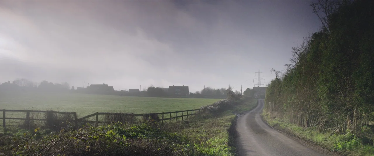 Bull (2021) movie still: establishing — A quiet rural road curves alongside a fenced green pasture under heavy clouds. In the distance, several…