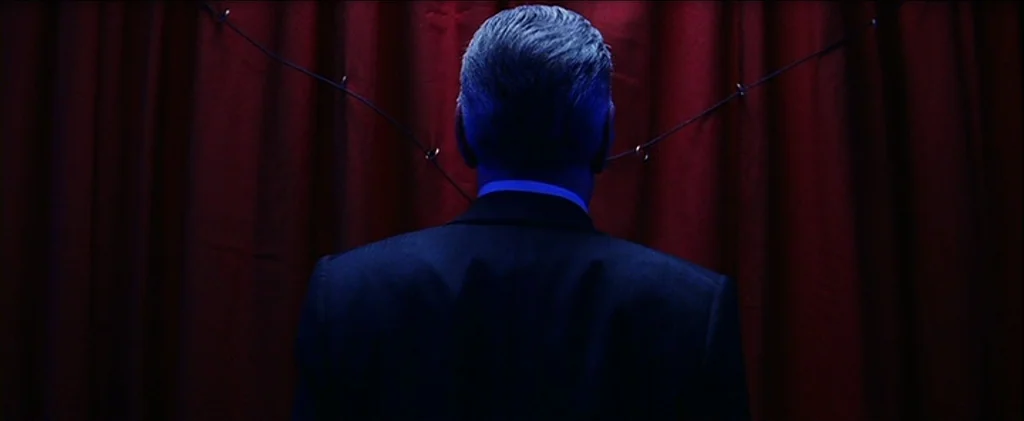 Magnolia (1999) movie still: medium — A dark, red curtain hangs behind a person standing in an interior space. The person in a dark suit faces the…