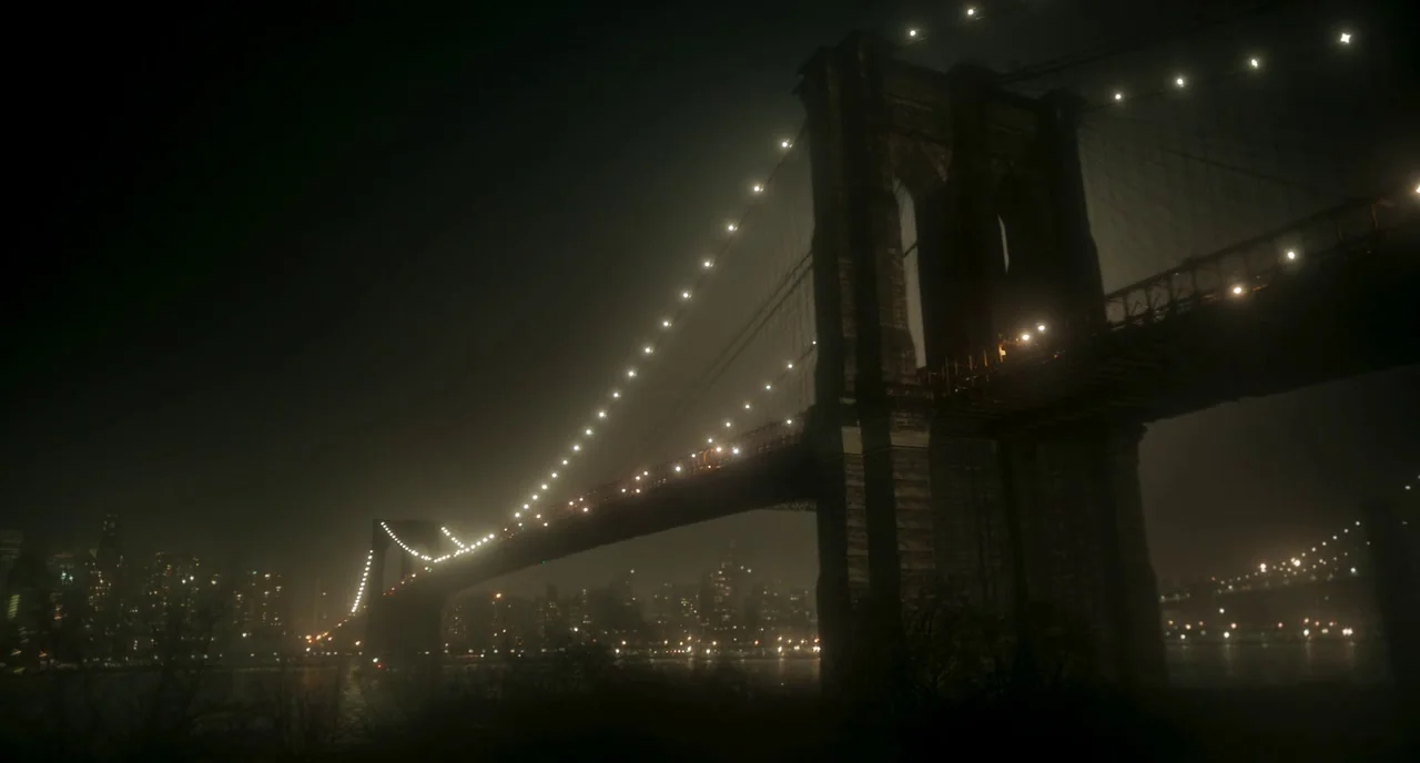 Motherless Brooklyn (2019) movie still: establishing — A massive suspension bridge looms over the dark river under a thick night haze. The city…
